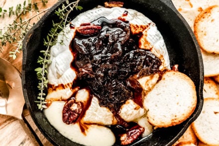 Overhead view of baked brie in a small cast-iron pan, topped with rich caramelized onions and pecans, served alongside toasted baguette slices on a wooden board with fresh thyme and a serving spoon