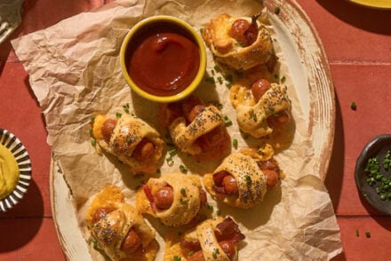 Overhead view of Bacon Pigs in a Blanket served on parchment paper and yellow plates, each wrapped in flaky crescent dough with crisp bacon and poppy seeds, accompanied by small bowls of ketchup and mustard for dipping, set on a warm red tile surface with drinks and a yellow gingham napkin for a cozy party presentation.