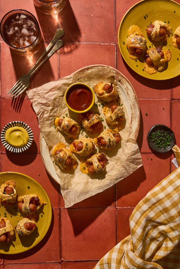 Overhead view of Bacon Pigs in a Blanket served on parchment paper and yellow plates, each wrapped in flaky crescent dough with crisp bacon and poppy seeds, accompanied by small bowls of ketchup and mustard for dipping, set on a warm red tile surface with drinks and a yellow gingham napkin for a cozy party presentation.