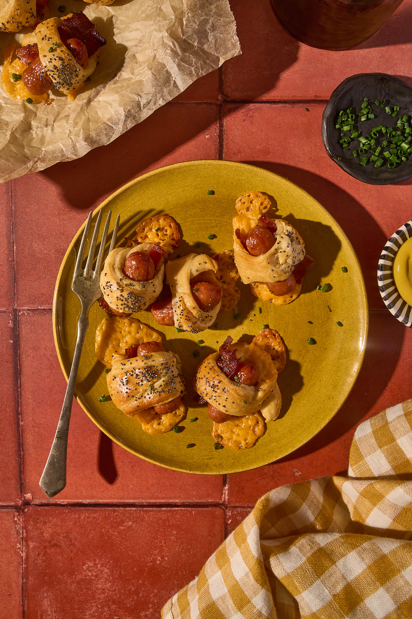 Overhead view of Bacon Pigs in a Blanket served on a mustard-yellow plate. Each mini sausage is wrapped in crispy bacon and golden crescent dough, sprinkled with poppy seeds and chives, with melted cheddar pooled underneath. A vintage fork rests on the plate, surrounded by terracotta tiles, a small dish of chopped chives, and a yellow gingham napkin for a cozy, casual presentation.