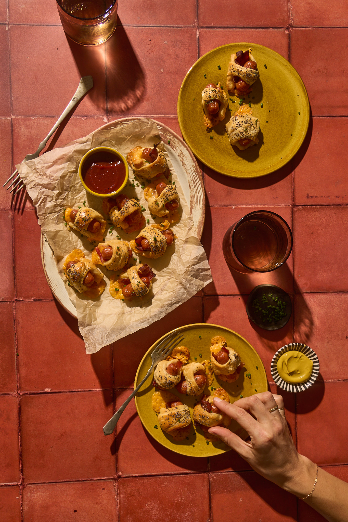 Top-down view of Bacon Pigs in a Blanket arranged on parchment paper and rustic yellow plates. Each mini cocktail sausage is wrapped in crispy bacon and golden-brown crescent dough, sprinkled with poppy seeds and chopped chives. A small bowl of ketchup sits at the center for dipping, alongside a dish of mustard. Two iced drinks and scattered forks complete the warm, inviting tabletop scene on terracotta tiles, creating a cozy party appetizer vibe.