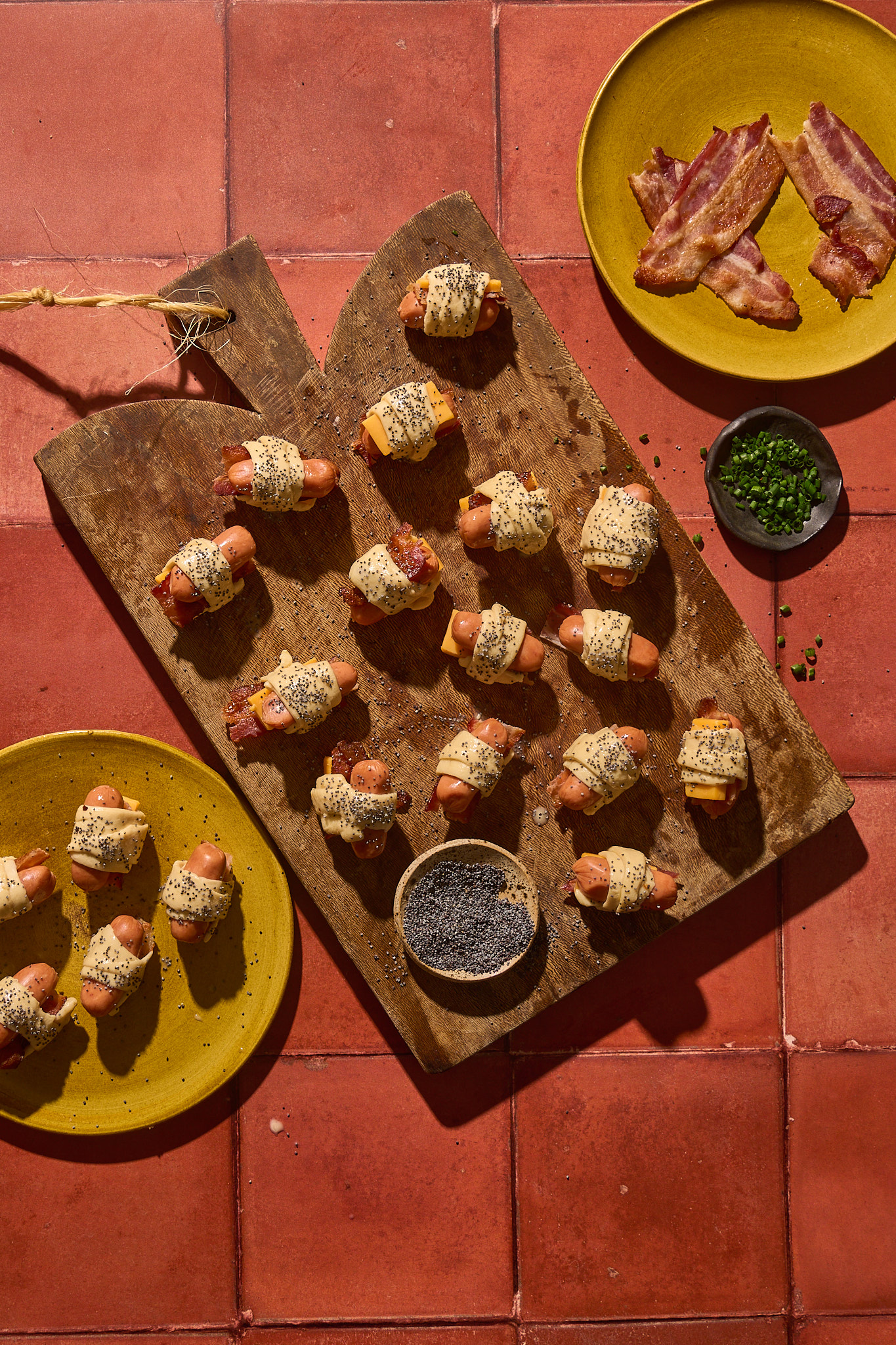 Overhead view of unbaked Bacon Pigs in a Blanket arranged on a rustic wooden board. Mini sausages are wrapped in crescent dough with strips of bacon and cheddar tucked inside, then brushed with egg wash and sprinkled generously with poppy seeds. A plate of extra cooked bacon and a small dish of chopped chives sit nearby on warm terracotta tiles, capturing the appetizer just before baking.