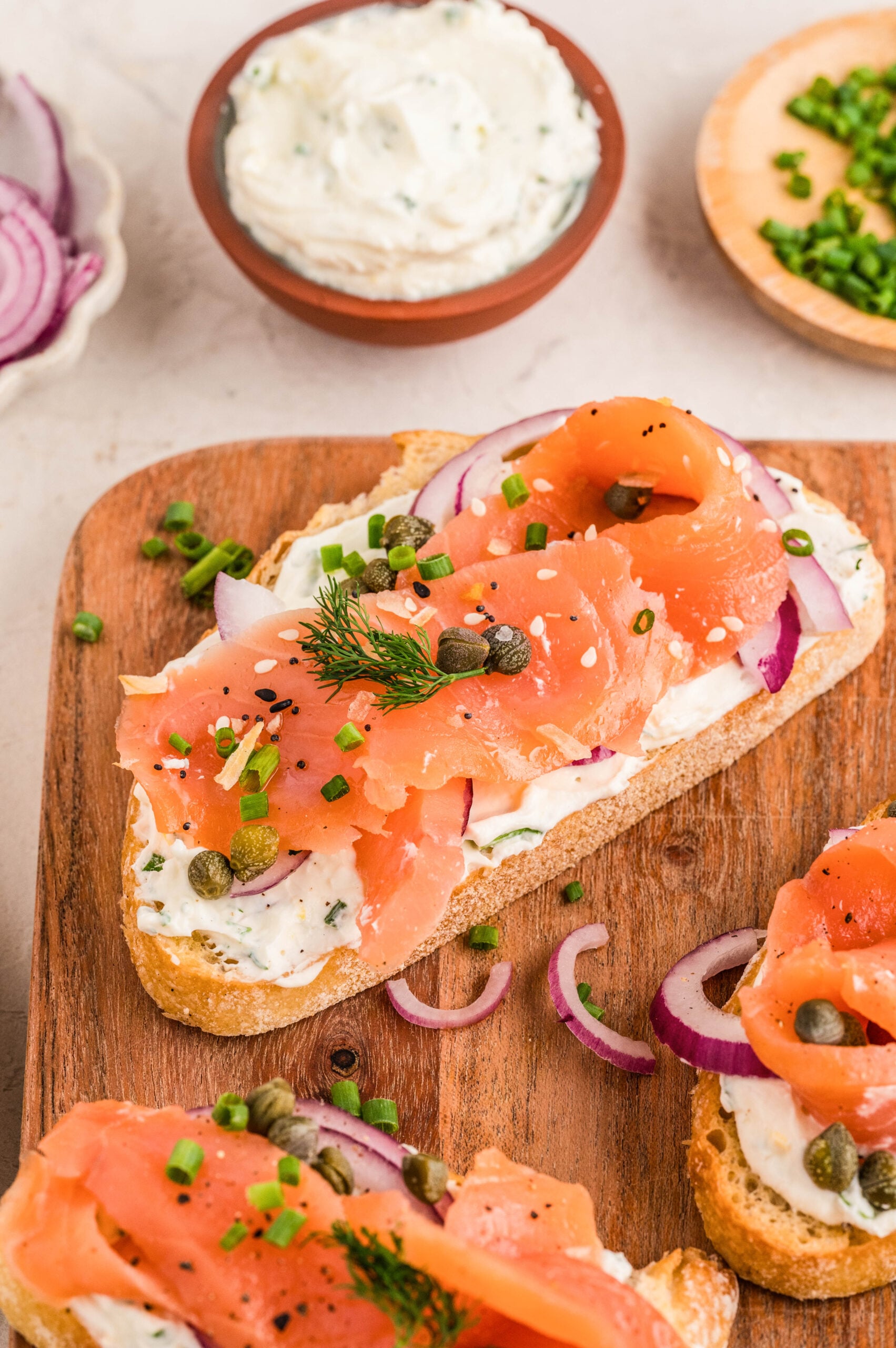 Close-up of Smoked Salmon Bruschetta on a wooden board, topped with lemon-herb cream cheese spread, smoked salmon ribbons, thin red onion slices, capers, fresh dill, and chopped chives, with extra spread and garnishes in the background