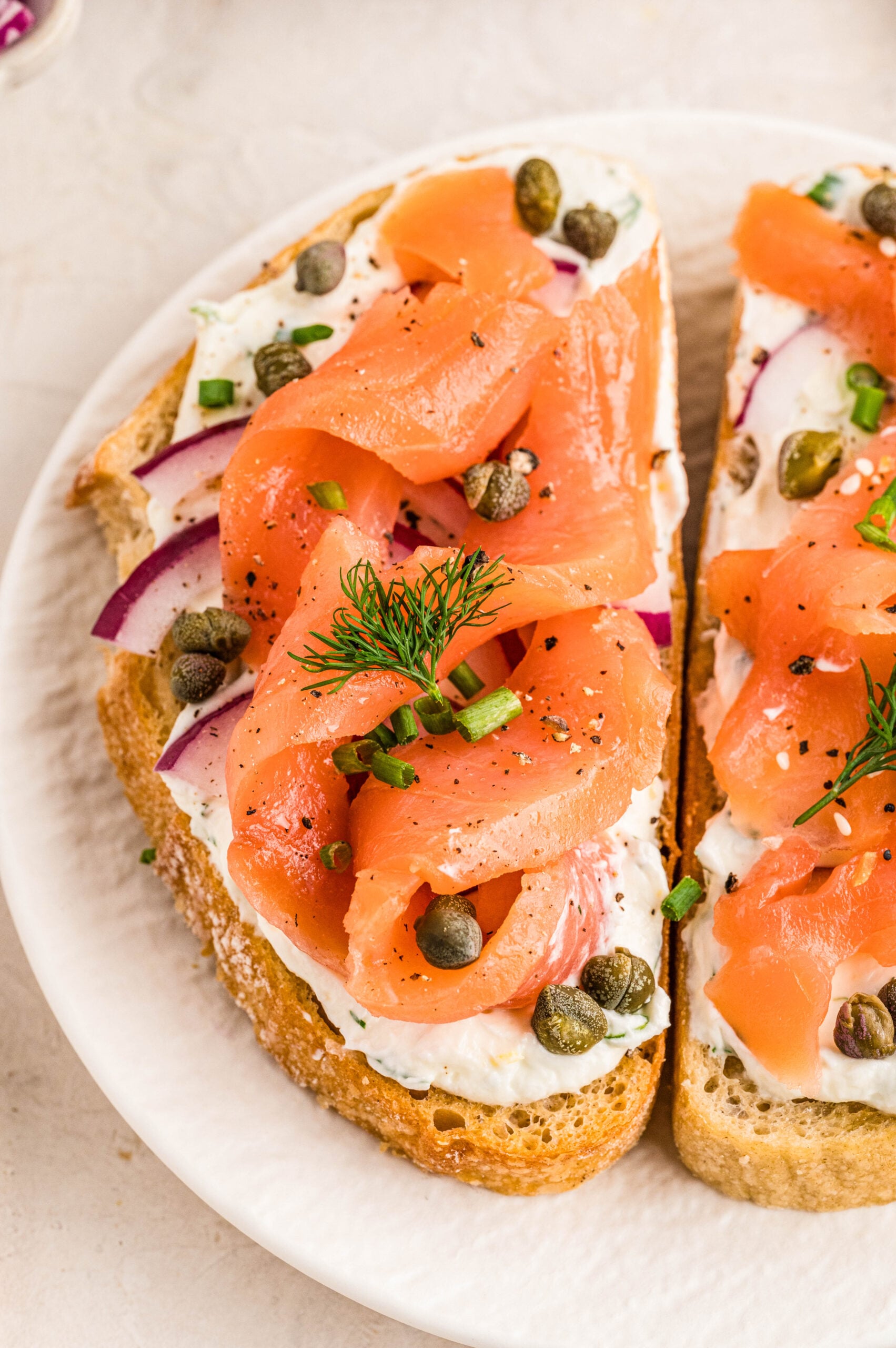 Close-up of Smoked Salmon Bruschetta on a white plate, featuring toasted bread topped with lemon-herb cream cheese, thin red onion slices, smoked salmon ribbons, capers, chopped chives, cracked pepper, and a sprig of fresh dill.