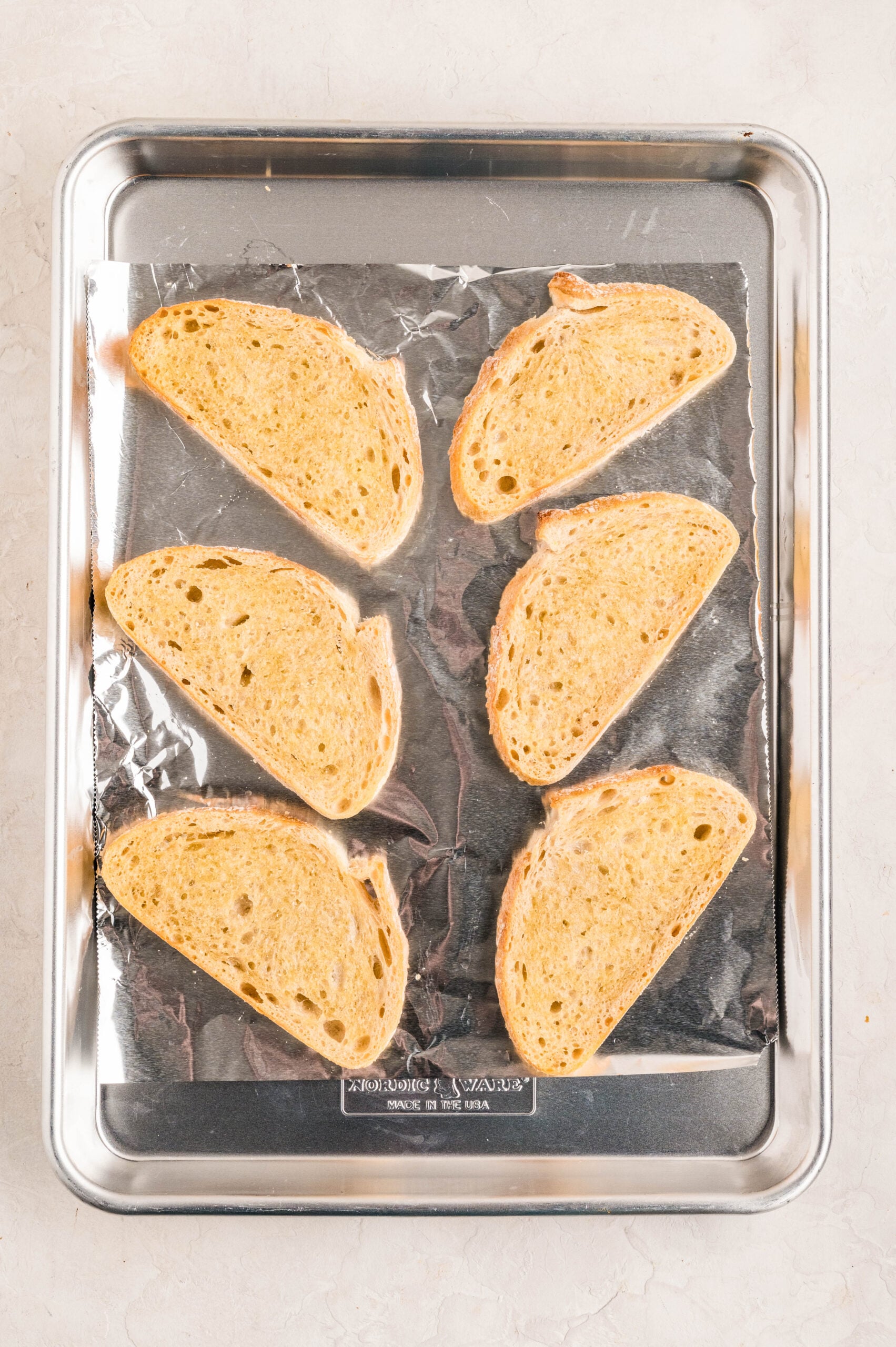 Six slices of crusty bread arranged on a foil-lined baking sheet, lightly toasted and ready to be topped for making Smoked Salmon Bruschetta