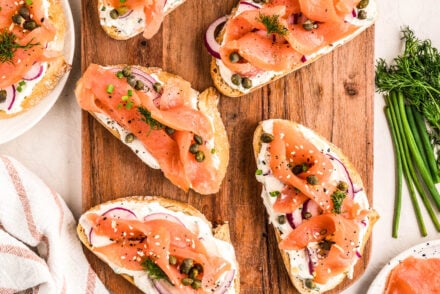 Smoked Salmon Bruschetta arranged on a wooden serving board, topped with a creamy lemon-herb spread, thin red onion slices, capers, fresh dill, and chives. Additional ingredients like sliced red onions, chives, dill, and smoked salmon surround the board for a fresh, vibrant presentation