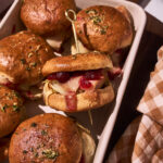 Close-up of baked Thanksgiving turkey leftover sliders in a white baking dish, with golden toasted rolls brushed with garlic herb butter, melted provolone cheese, stuffing, and cranberry sauce visible inside one slider secured with a toothpick.