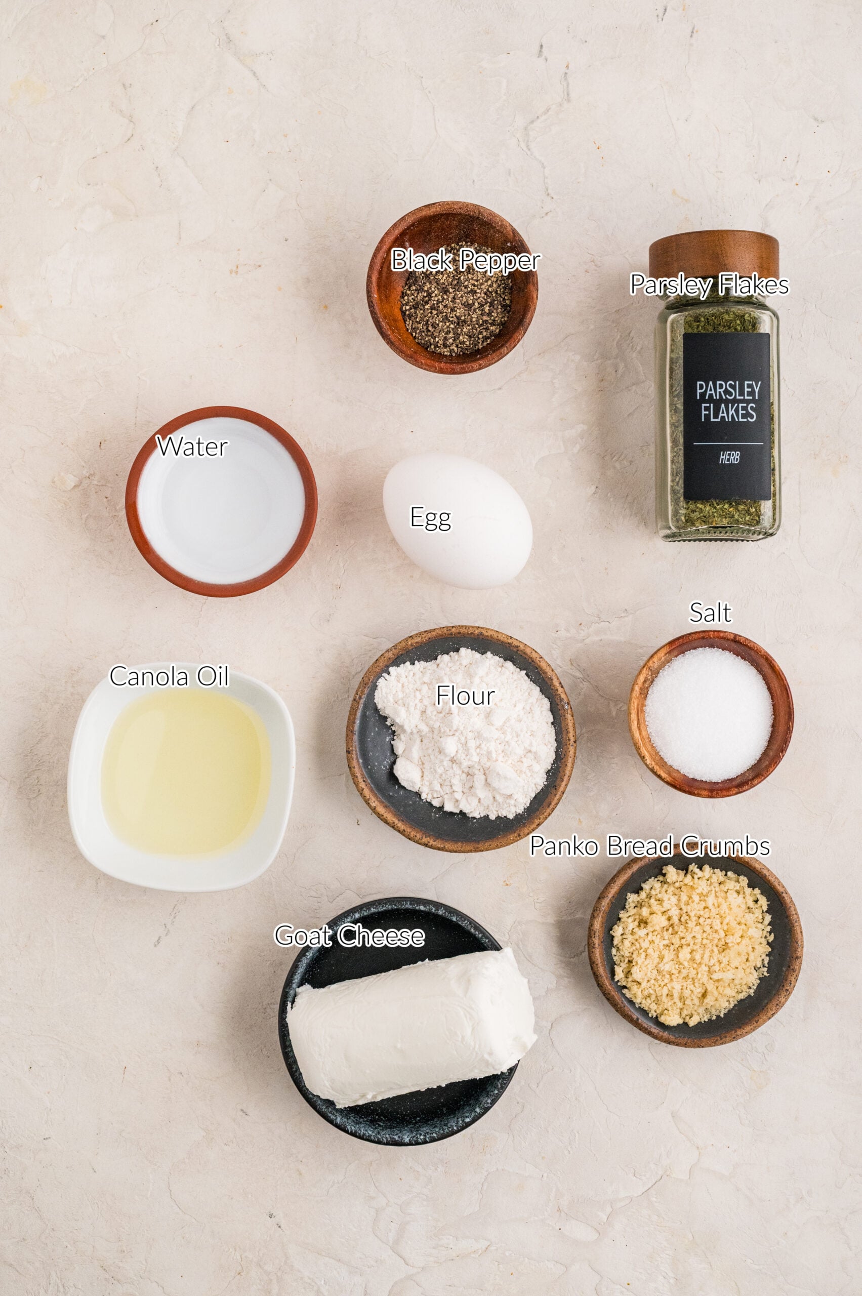 Overhead image of ingredients for fried goat cheese balls arranged neatly on a light surface, including a log of goat cheese, flour, panko breadcrumbs, salt, black pepper, parsley flakes, an egg, water, and canola oil in small bowls, each labeled for reference.
