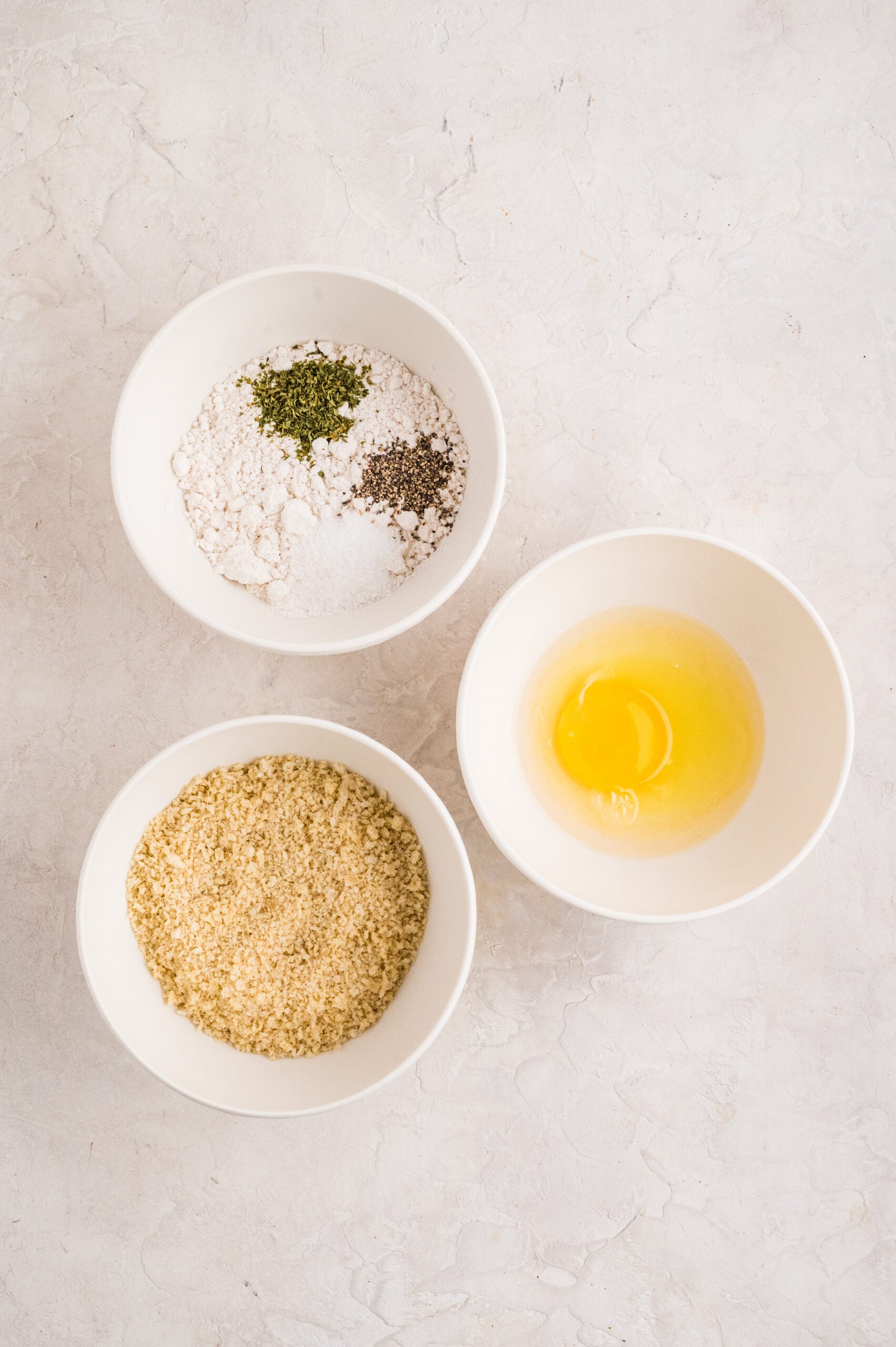 Overhead view of three bowls showing the dredging station for fried goat cheese balls: one bowl with flour mixed with parsley, salt, and black pepper, a second bowl with a cracked egg for the egg wash, and a third filled with panko breadcrumbs, all arranged on a light textured surface.