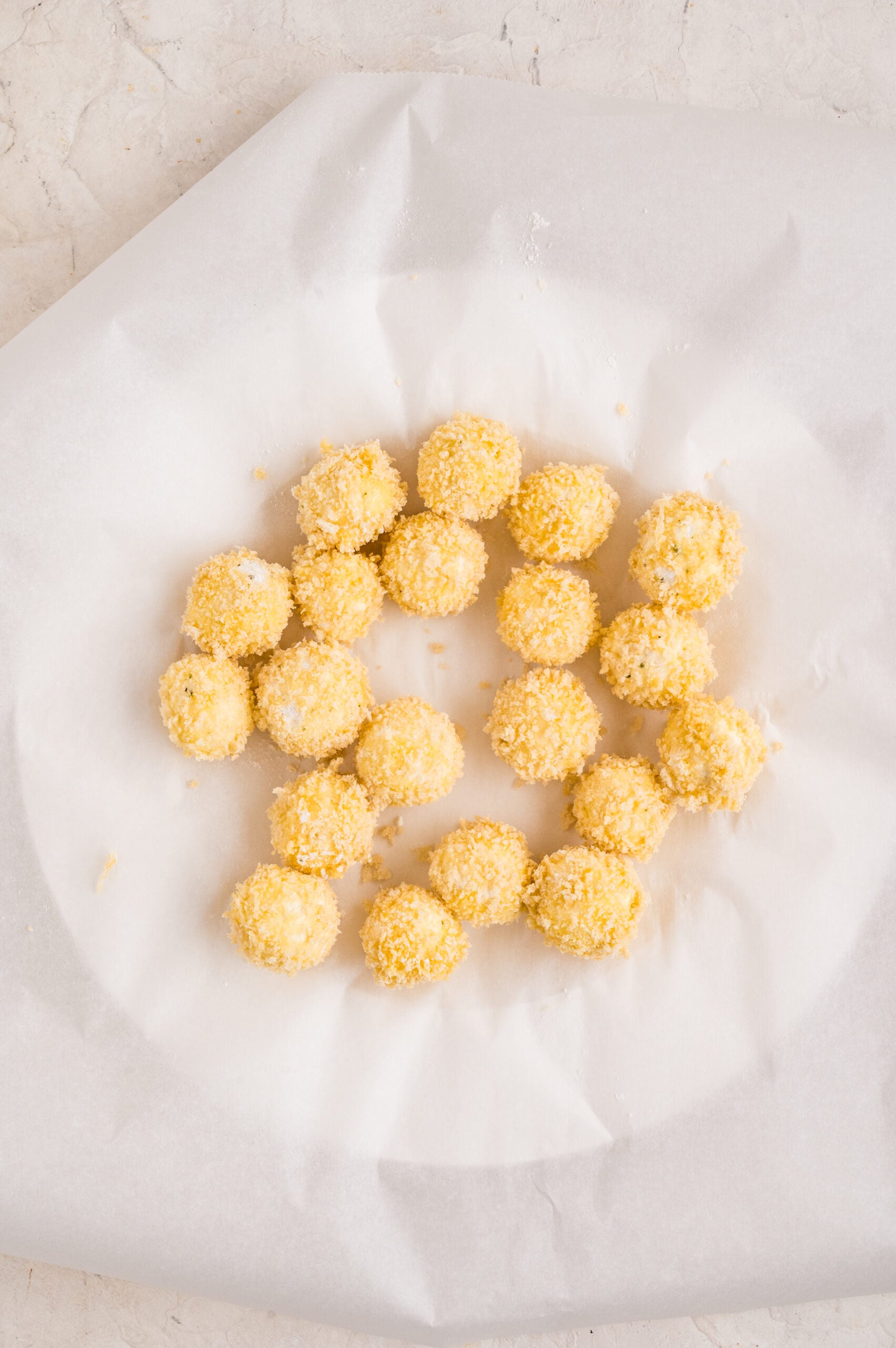 Overhead image of uncooked fried goat cheese balls arranged in a circle on parchment paper, each piece coated in a light golden layer of panko breadcrumbs, ready to be frozen before frying.
