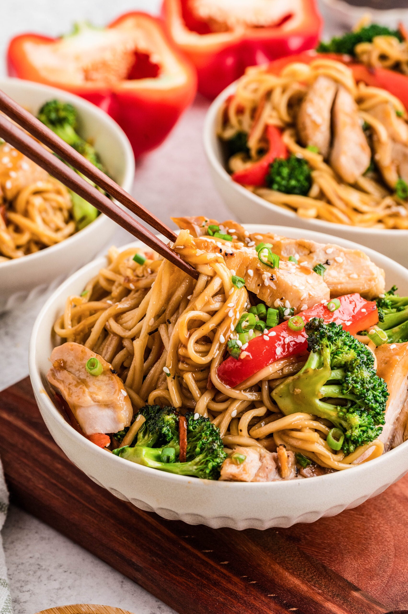 A bowl of Chicken Teriyaki Bowls loaded with saucy lo mein noodles, sliced chicken, broccoli florets, and red bell pepper strips, topped with sesame seeds and green onions, with chopsticks lifting a bite and additional bowls and bell peppers in the background.