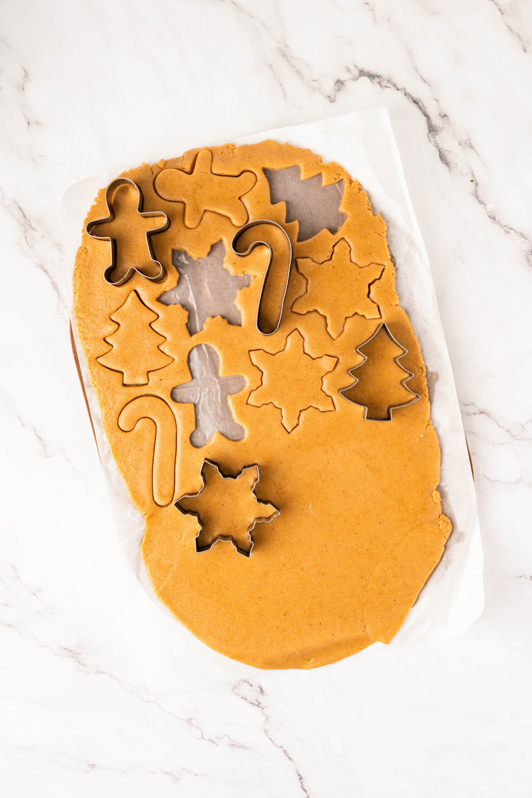 Rolled-out gingerbread cookie dough on parchment paper with metal cookie cutters cutting stars, trees, candy canes, and gingerbread people, preparing frosted gingerbread cookies for holiday baking.