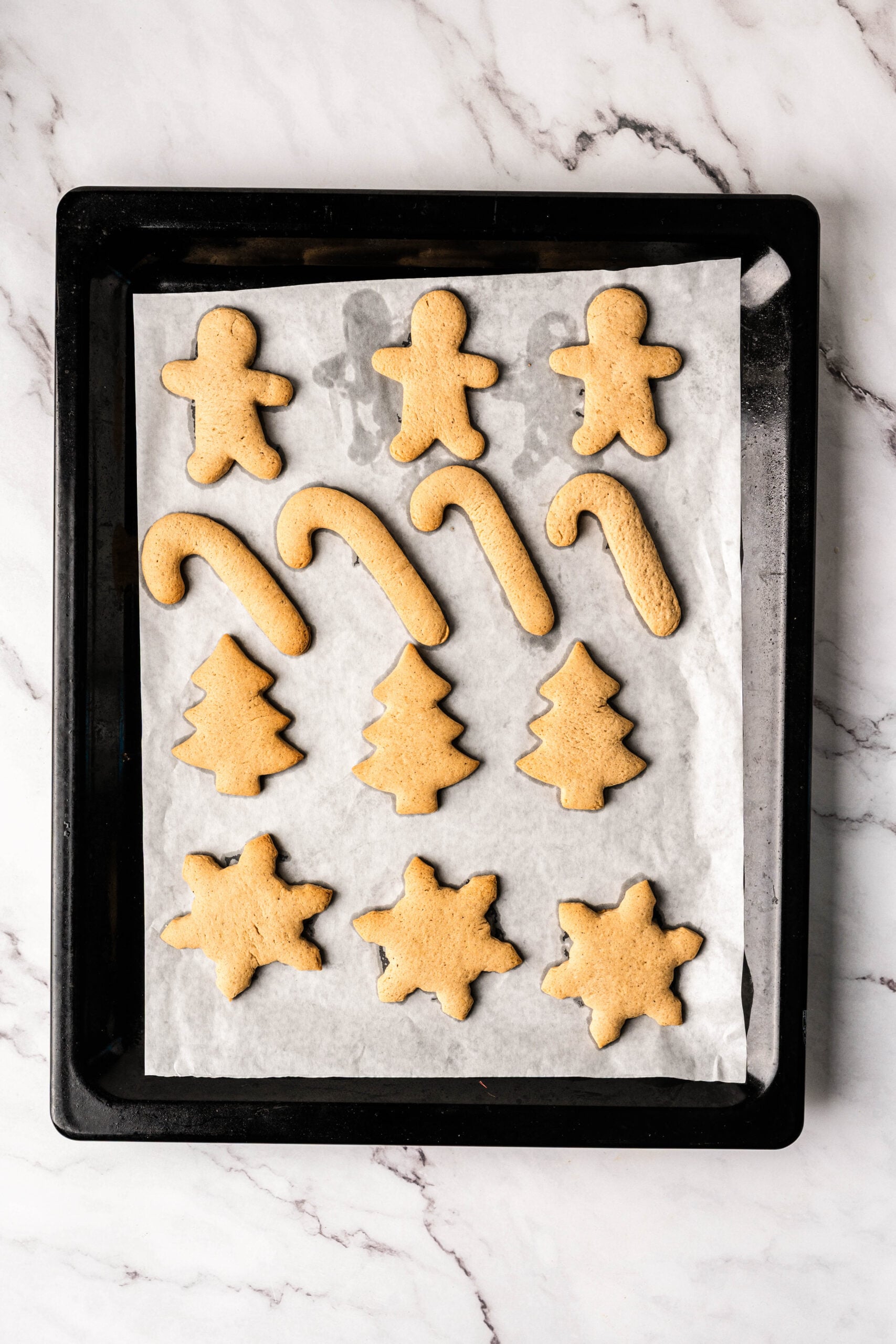 Baked gingerbread cookies shaped like gingerbread people, candy canes, Christmas trees, and stars cooling on a parchment-lined baking sheet, ready to be decorated as frosted gingerbread cookies.