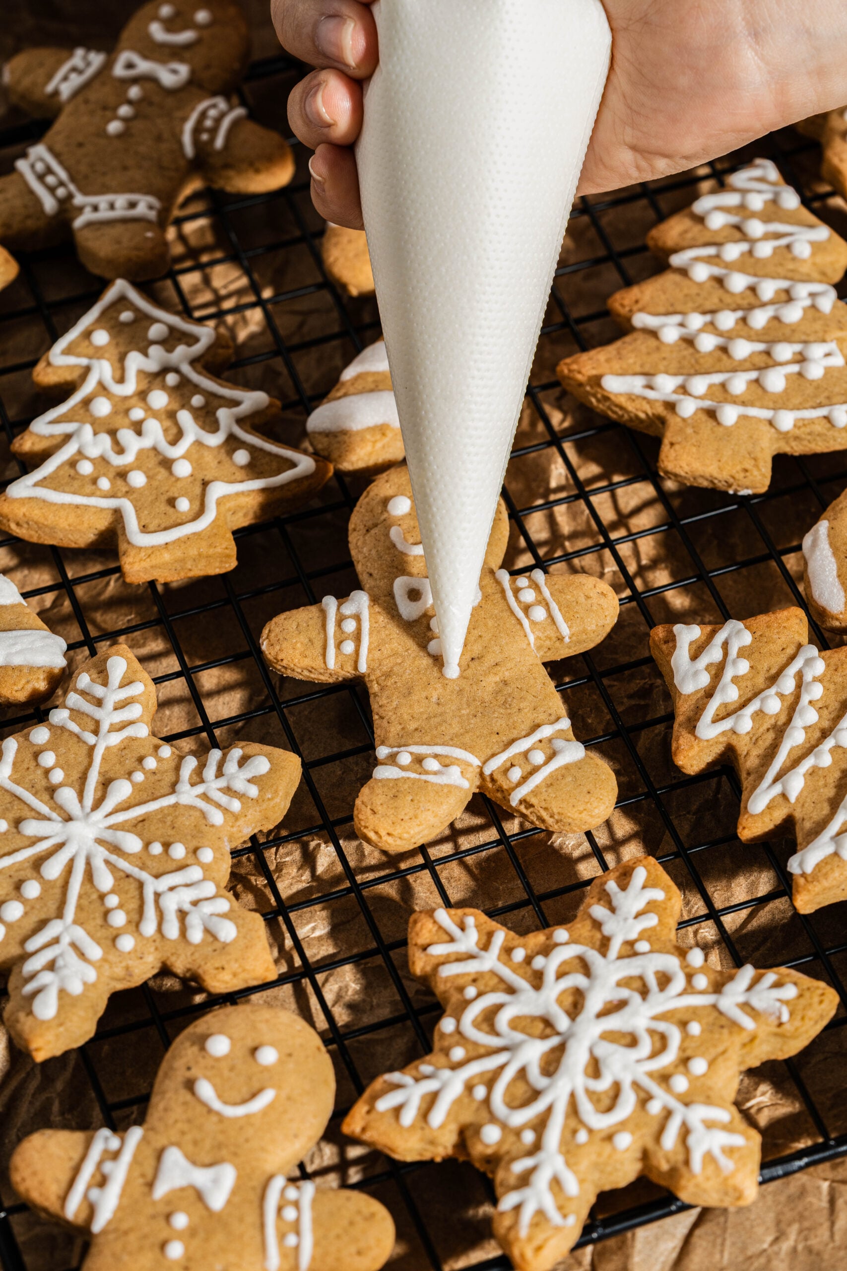 Hand piping white icing onto gingerbread people, snowflakes, and Christmas tree cookies on a cooling rack, decorating frosted gingerbread cookies with detailed holiday designs.