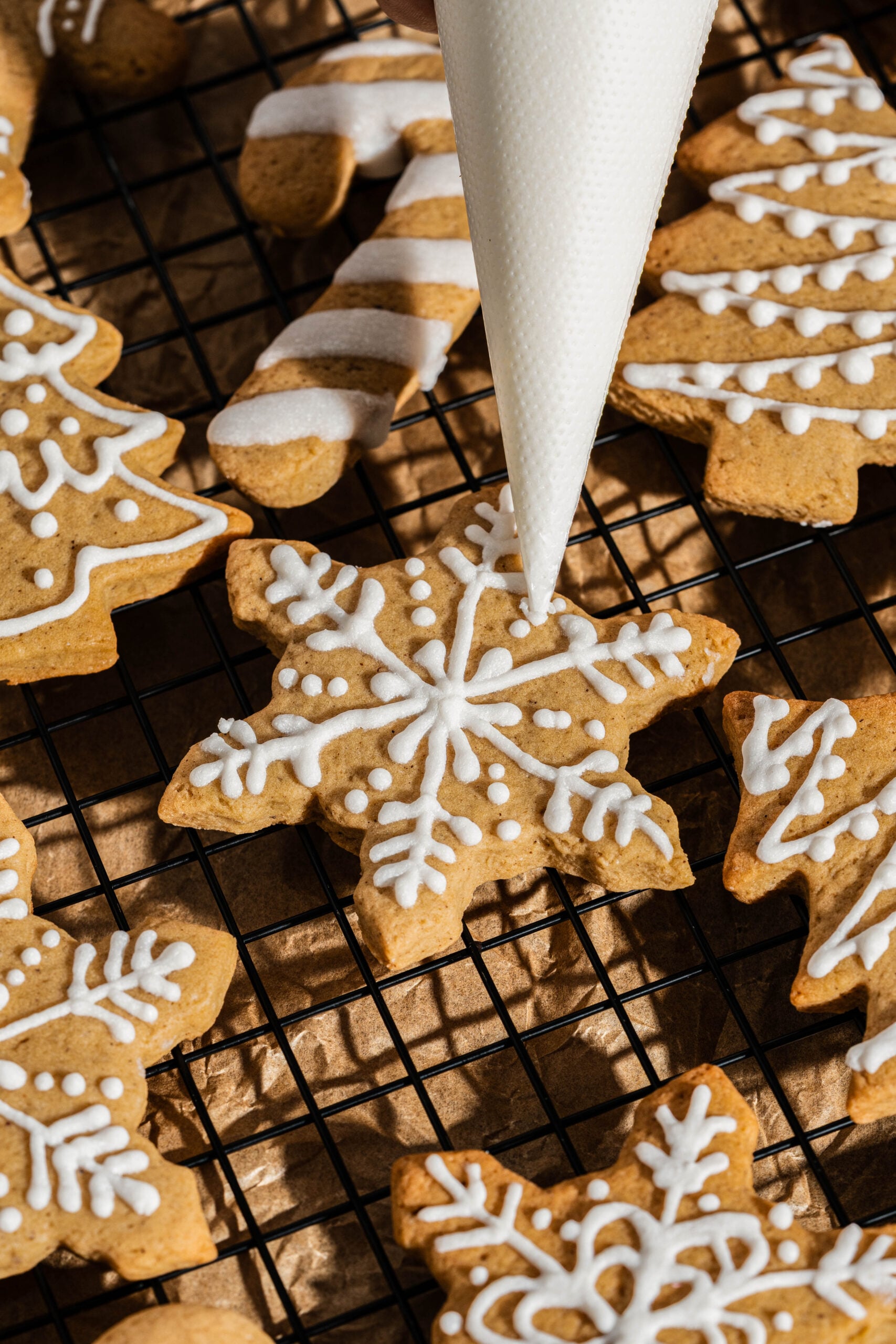 Hand piping white icing onto gingerbread people, snowflakes, and Christmas tree cookies on a cooling rack, decorating frosted gingerbread cookies with detailed holiday designs.