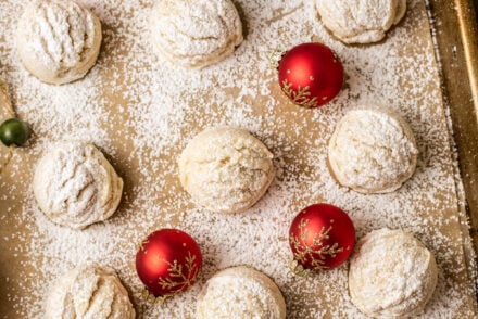 Snowball cookies without nuts arranged on a parchment-lined baking sheet, each dusted heavily with powdered sugar to resemble fresh snow. The cookies are surrounded by festive holiday decorations—including red ornaments, candy canes, a miniature Christmas tree, and gingerbread garland—creating a cozy, winter-themed presentation.