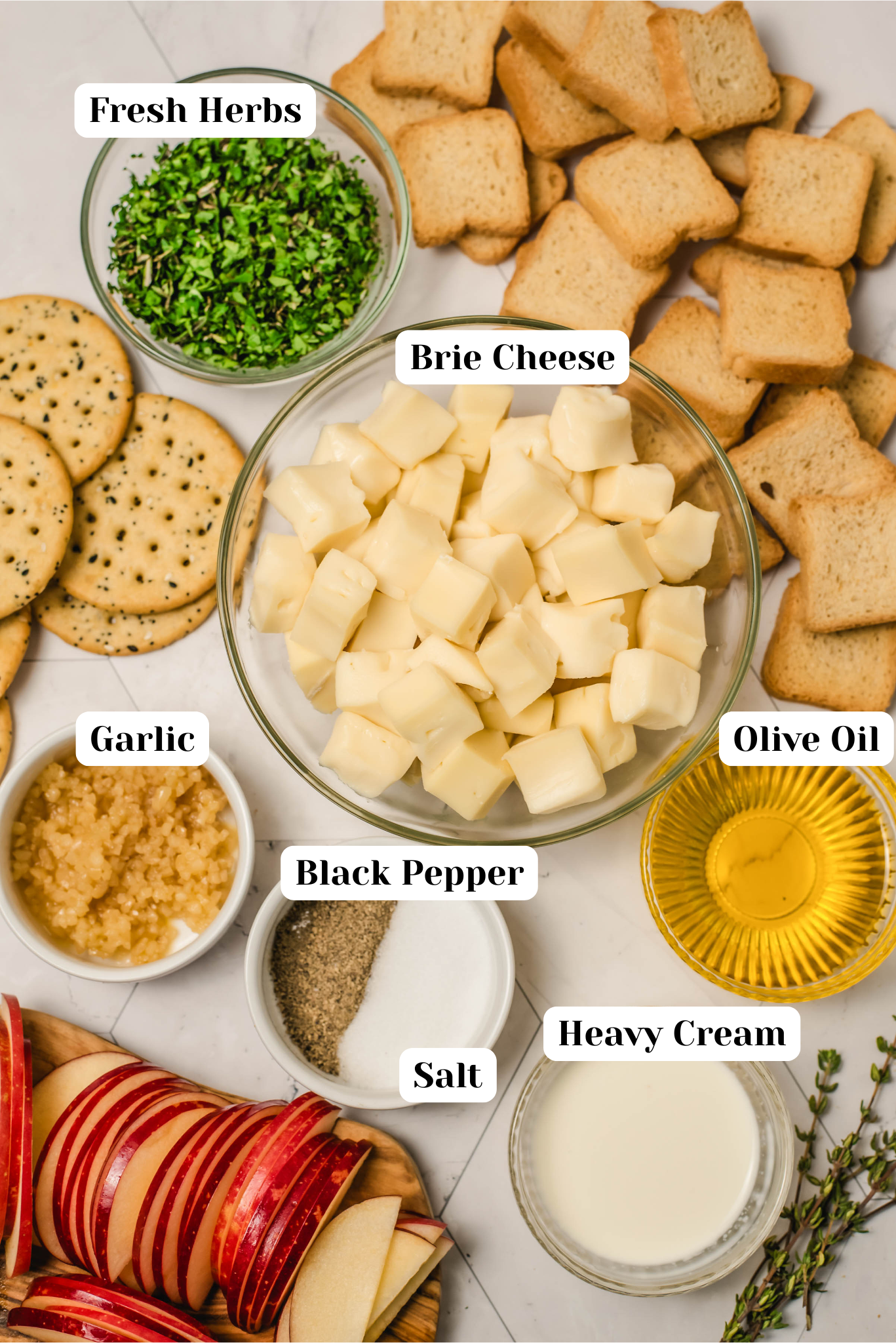 Overhead ingredient layout for whipped brie, featuring cubed brie cheese, fresh herbs, garlic, olive oil, heavy cream, salt, black pepper, crostini, crackers, and sliced apples on a light marble surface.