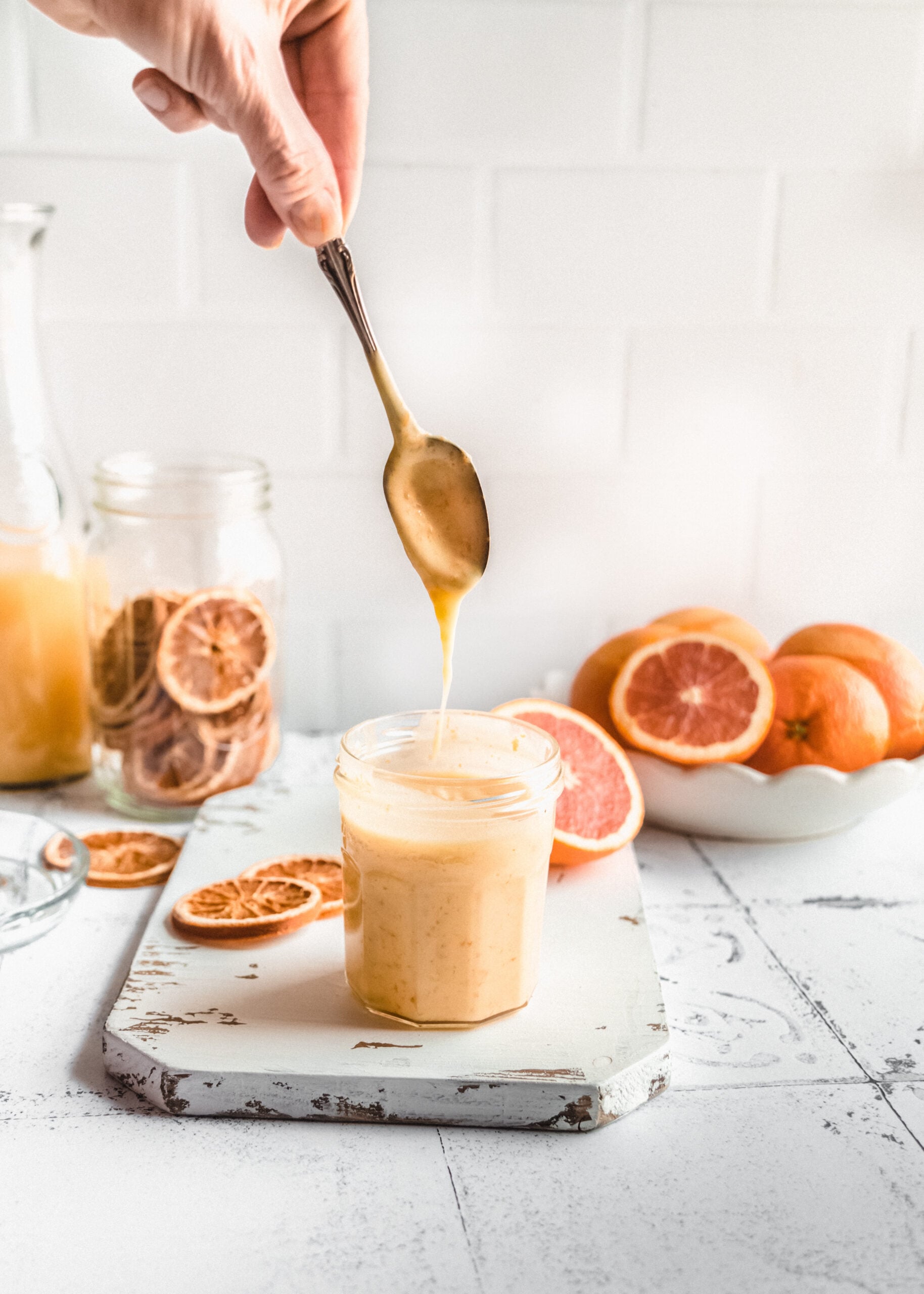 Spoon dripping creamy homemade orange curd into a small glass jar on a white marble surface, surrounded by fresh oranges, sliced citrus, and jars of orange juice for a bright orange curd recipe scene.