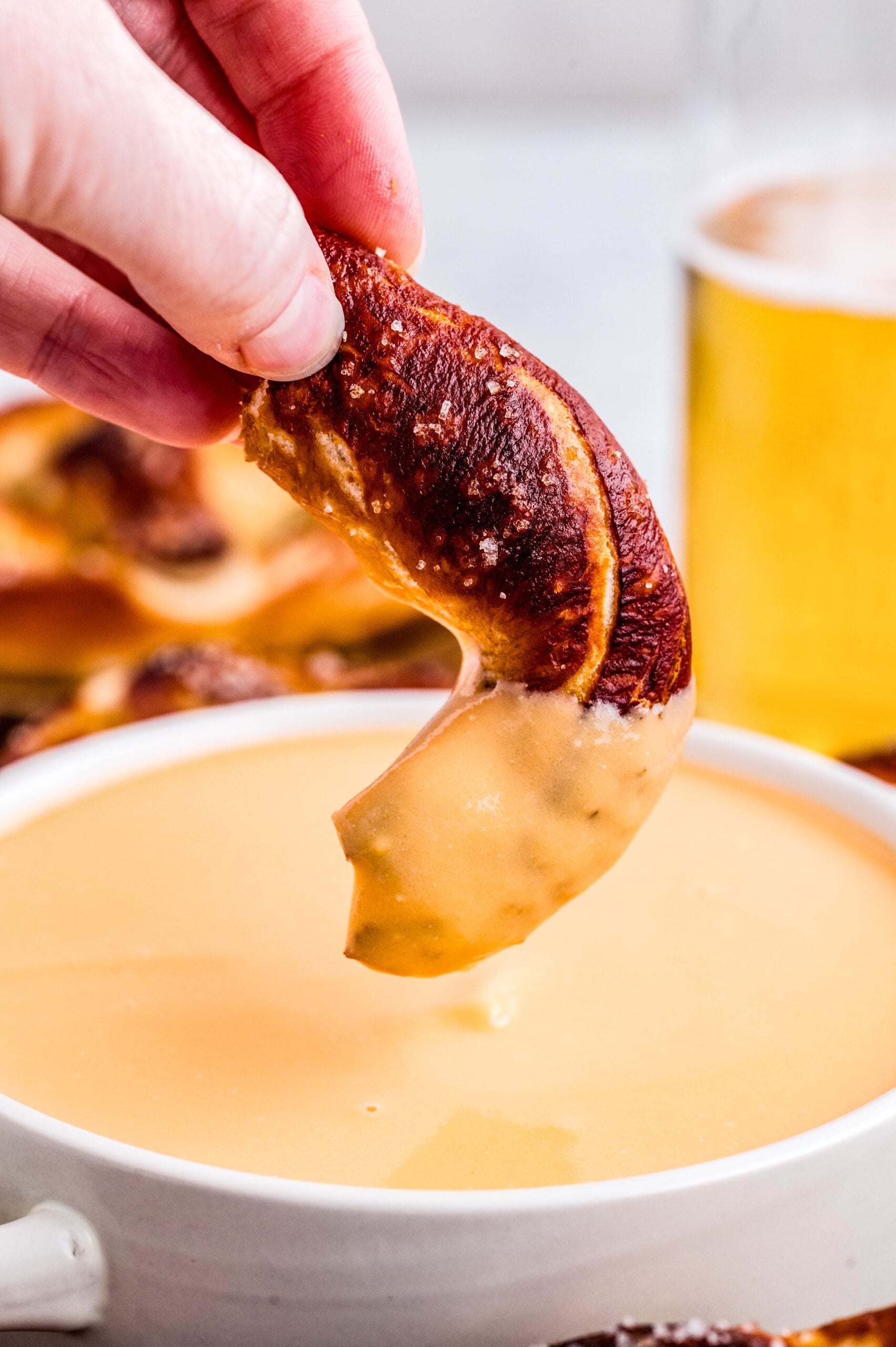 Hand dipping a soft salted pretzel into creamy Three-Ingredient Beer Cheese Dip, with melted cheese stretching back into the bowl.