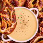 Overhead view of Three-Ingredient Beer Cheese Dip in a white bowl, sprinkled with cracked black pepper and surrounded by soft salted pretzels on a wooden board.