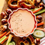 Overhead view of Three-Ingredient Beer Cheese Dip sprinkled with black pepper, surrounded by soft pretzels, pretzel bites, and fresh vegetables on a wooden board.