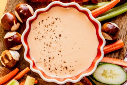 Overhead view of Three-Ingredient Beer Cheese Dip sprinkled with black pepper, surrounded by soft pretzels, pretzel bites, and fresh vegetables on a wooden board.