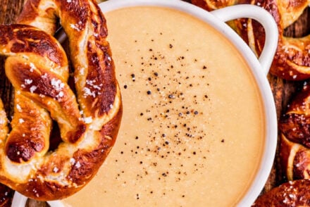 Overhead view of Three-Ingredient Beer Cheese Dip in a white bowl, sprinkled with cracked black pepper and surrounded by soft salted pretzels on a wooden board.
