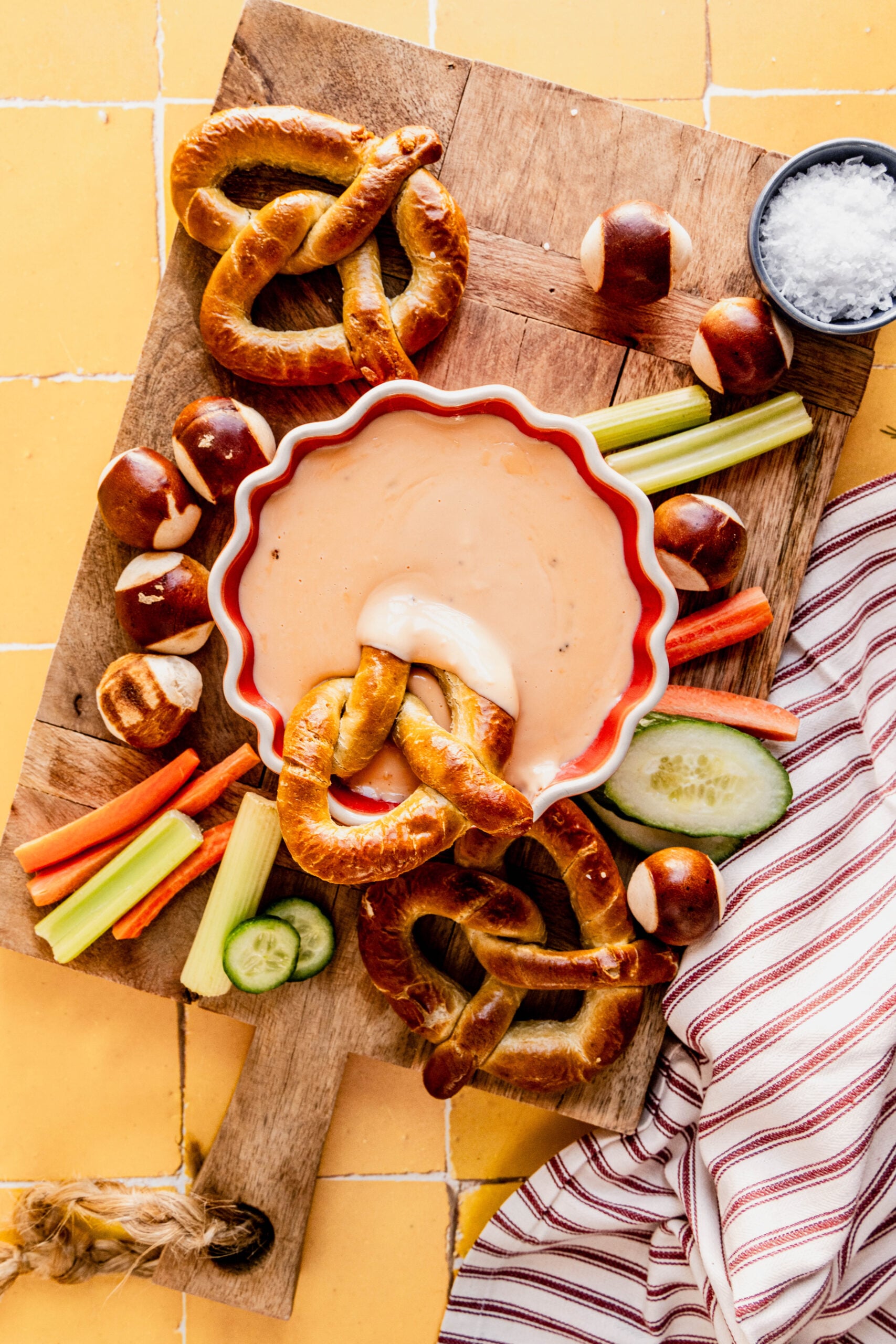 Overhead view of Three-Ingredient Beer Cheese Dip with a soft pretzel dipped into the creamy cheese, surrounded by pretzels, pretzel bites, and fresh vegetables on a wooden board.