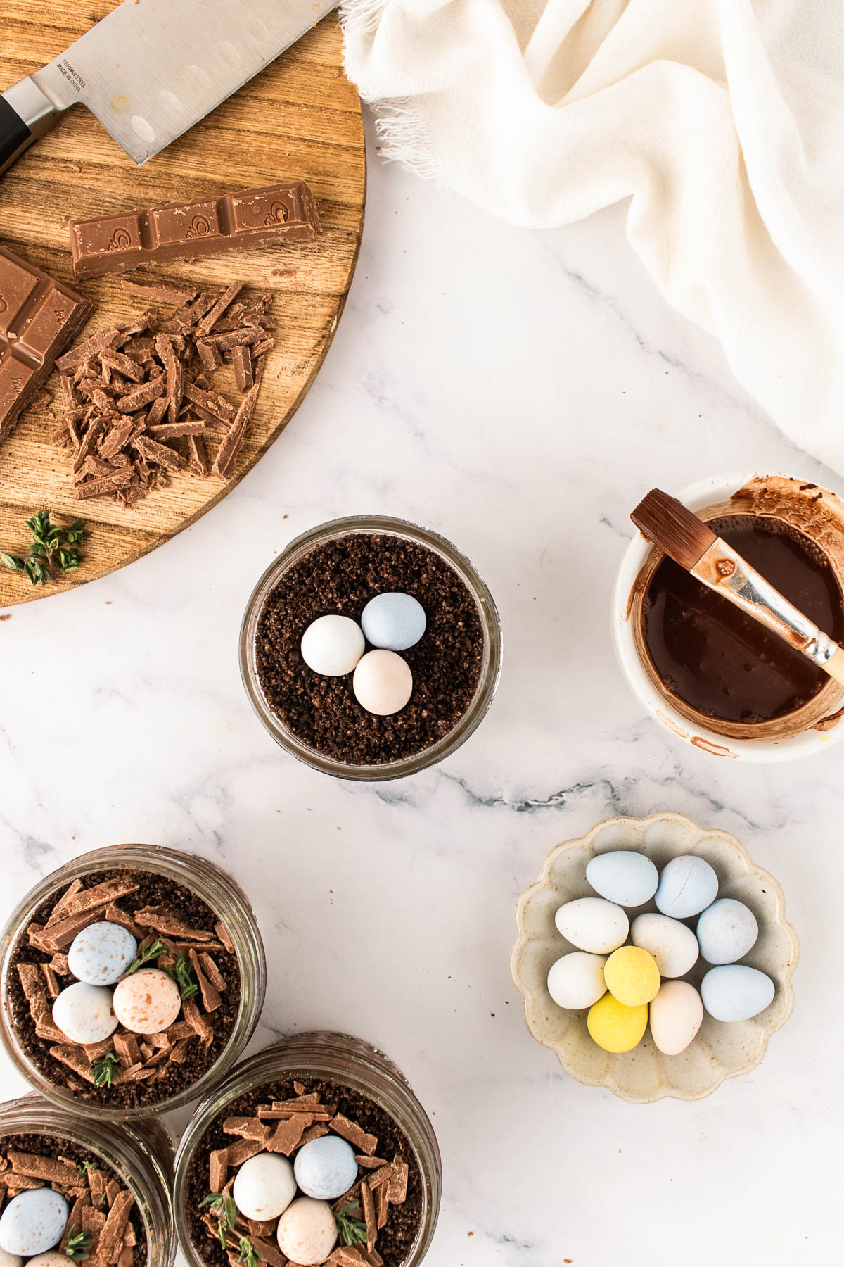 Overhead view of assembling Easter dirt cups with chocolate pudding, Oreo crumb “dirt,” chopped chocolate nests, and pastel speckled mini eggs, styled on a white marble surface with spring decor and baking tools.