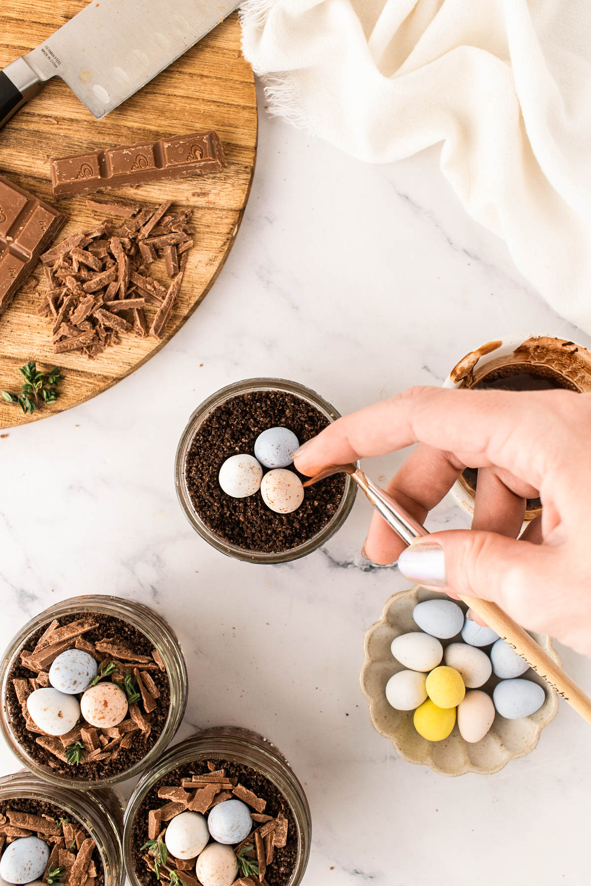 Speckling pastel mini chocolate eggs with cocoa powder while decorating Easter dirt cups, shown overhead with chocolate pudding cups, Oreo crumb “dirt,” chopped chocolate nests, and spring Easter styling on a white marble surface.