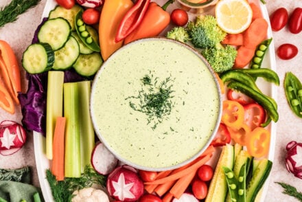 Overhead view of a vibrant vegetable platter featuring creamy green goddess dressing dip in the center, surrounded by fresh vegetables including cucumbers, carrots, snap peas, radishes, bell peppers, cherry tomatoes, broccoli, cauliflower, celery, and fresh herbs on a light neutral background.