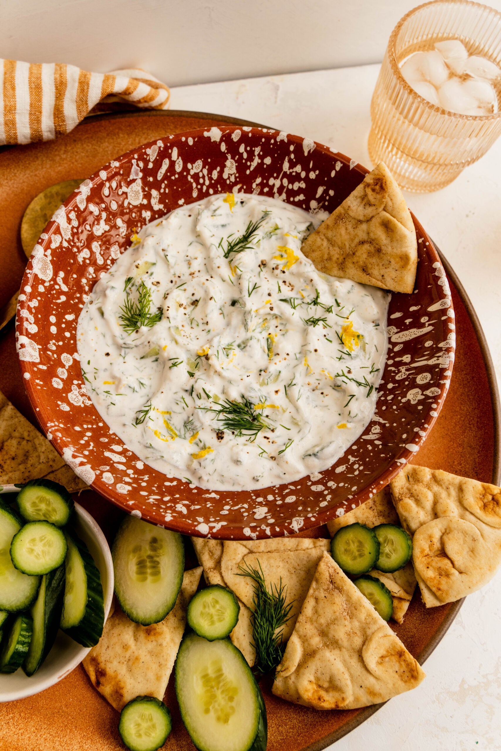 Overhead view of healthy tzatziki sauce served in a ceramic bowl, garnished with fresh dill, lemon zest, and cracked black pepper, surrounded by pita chips and sliced cucumbers on a warm-toned serving board.