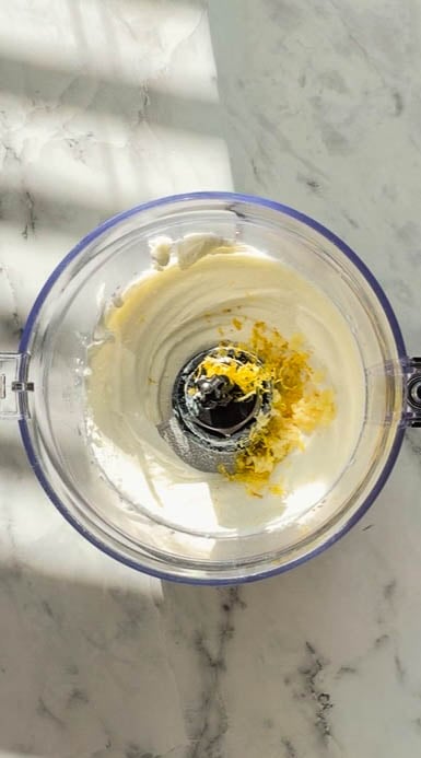Overhead view of healthy tzatziki sauce being blended in a food processor, with creamy yogurt and cottage cheese base topped with fresh lemon zest on a white marble surface.