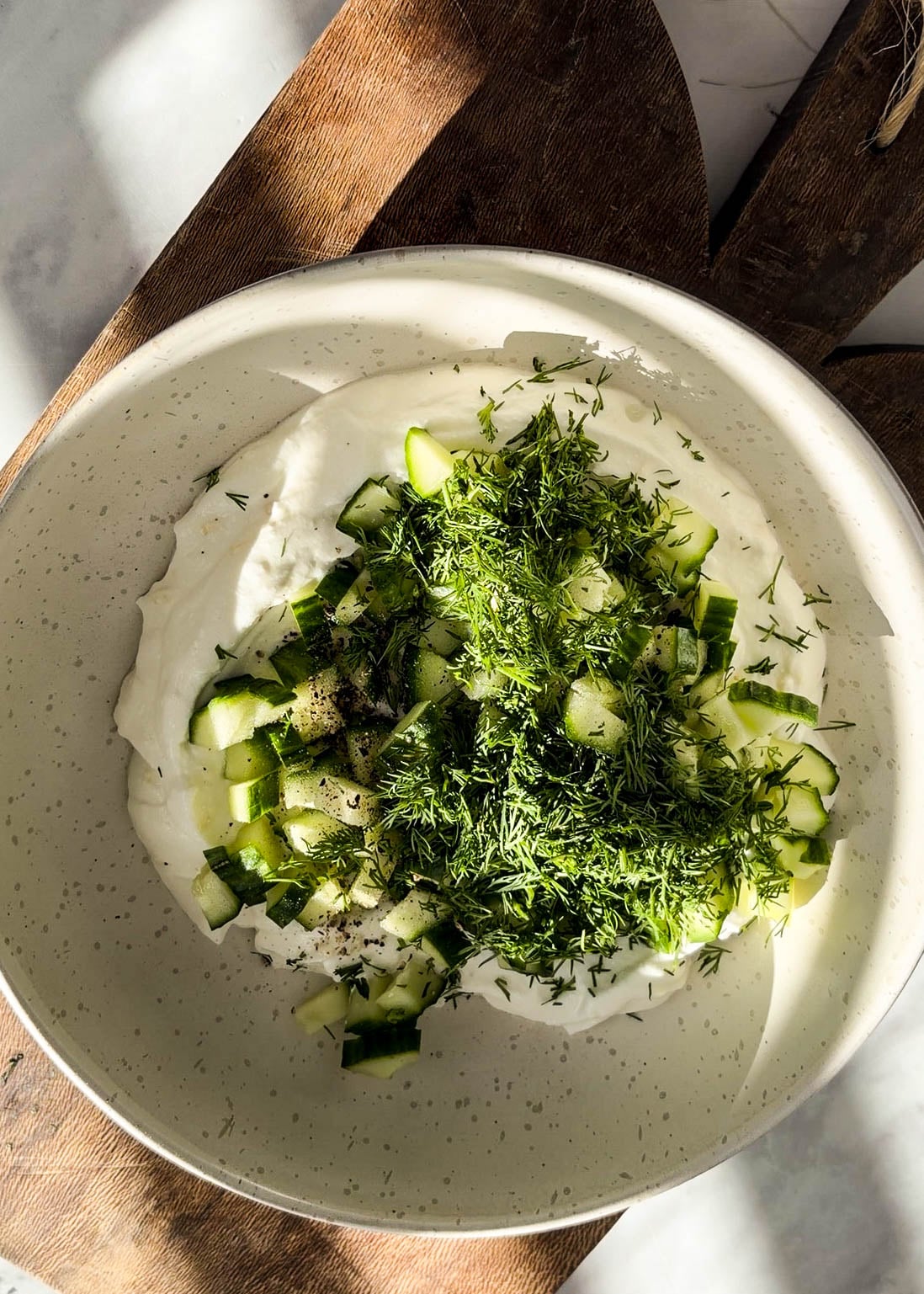 Overhead view of healthy tzatziki sauce with creamy yogurt base topped with freshly chopped cucumbers and fresh dill in a ceramic bowl on a wooden board over a white marble surface.