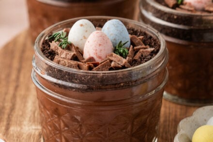Easter dirt cups layered with rich chocolate pudding and Oreo crumb “soil,” topped with delicate chocolate nests, speckled pastel mini eggs, and fresh thyme sprigs, served in clear glass jars on a wooden board.