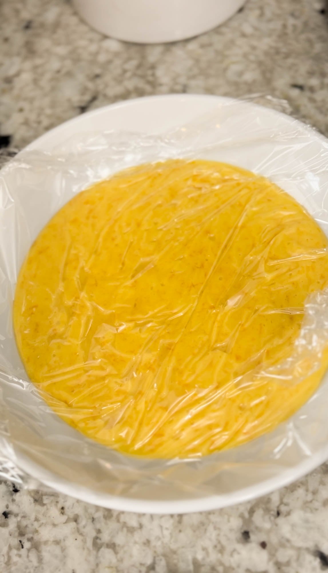 Freshly made orange curd cooling in a white bowl, covered with plastic wrap directly on the surface to prevent a skin from forming, shown during an orange curd recipe.