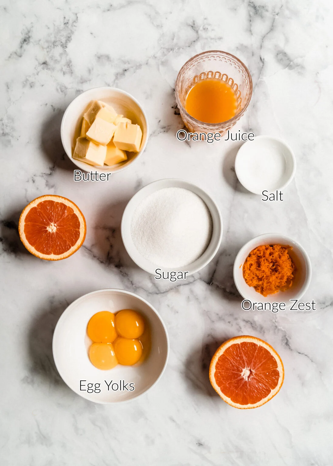 Overhead view of orange curd recipe ingredients arranged on a marble surface, including egg yolks, sugar, fresh orange juice, orange zest, butter, salt, and halved oranges.