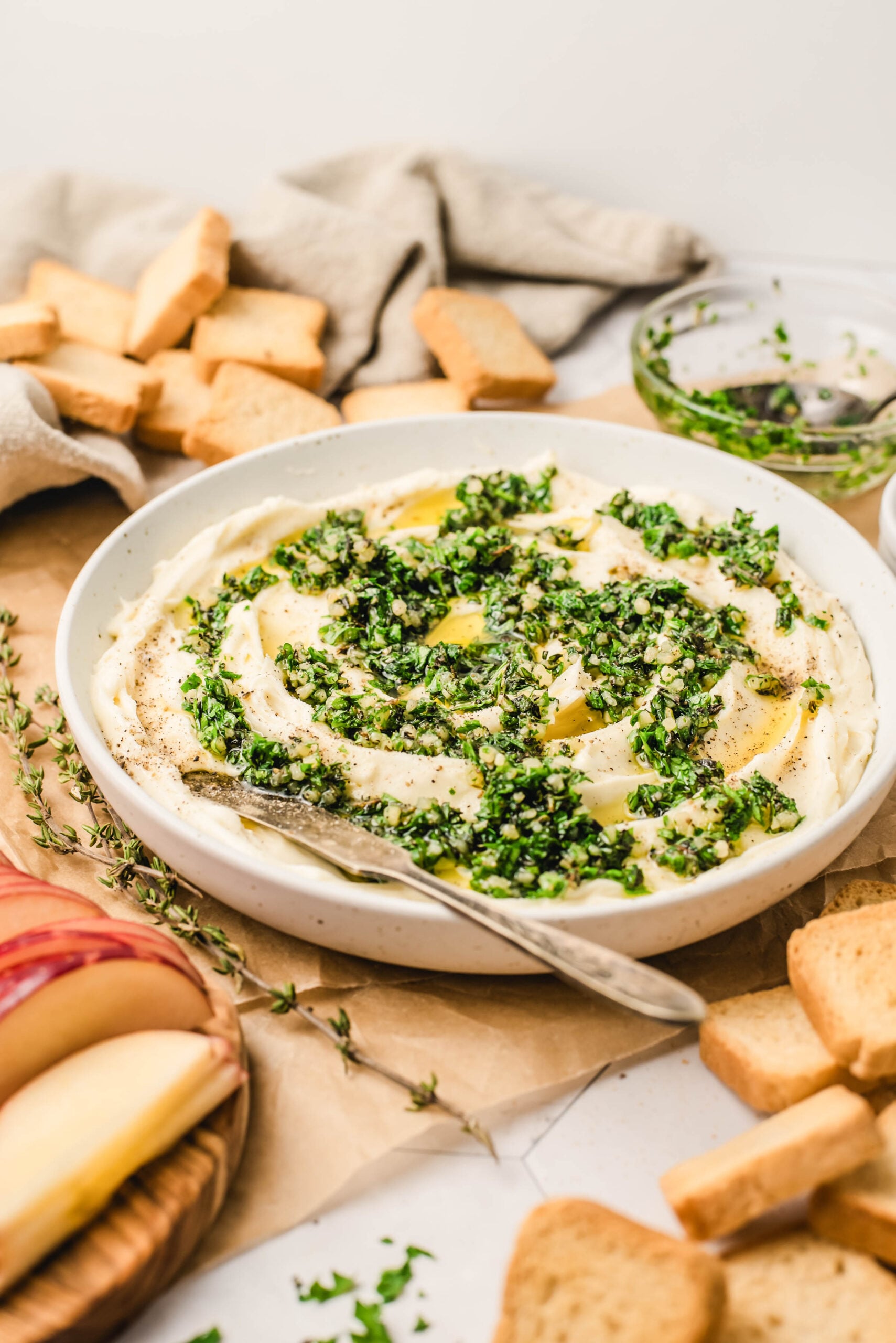 Whipped brie spread topped with chopped fresh herbs and olive oil in a shallow bowl, styled overhead with crostini, sliced apples, and a small spreading knife on a light neutral surface.