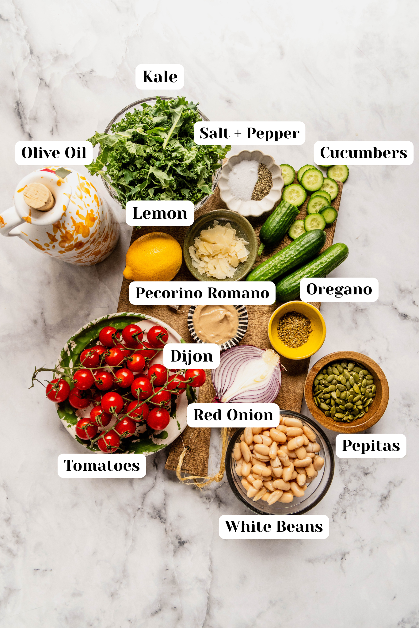Overhead ingredient shot for kale and cannellini bean salad featuring chopped kale, Persian cucumbers, grape tomatoes, cannellini beans, sliced red onion, pepitas, Pecorino Romano, lemon, Dijon mustard, olive oil, oregano, and salt and pepper arranged on a wood board over a white marble background.