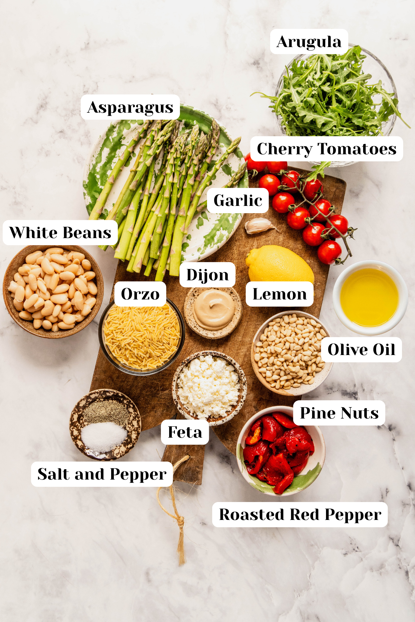“Overhead view of ingredients for asparagus orzo salad including fresh asparagus, orzo pasta, cherry tomatoes, arugula, white beans, roasted red peppers, feta cheese, pine nuts, garlic, lemon, Dijon mustard, olive oil, and salt and pepper arranged on a wooden cutting board over a white marble background.”