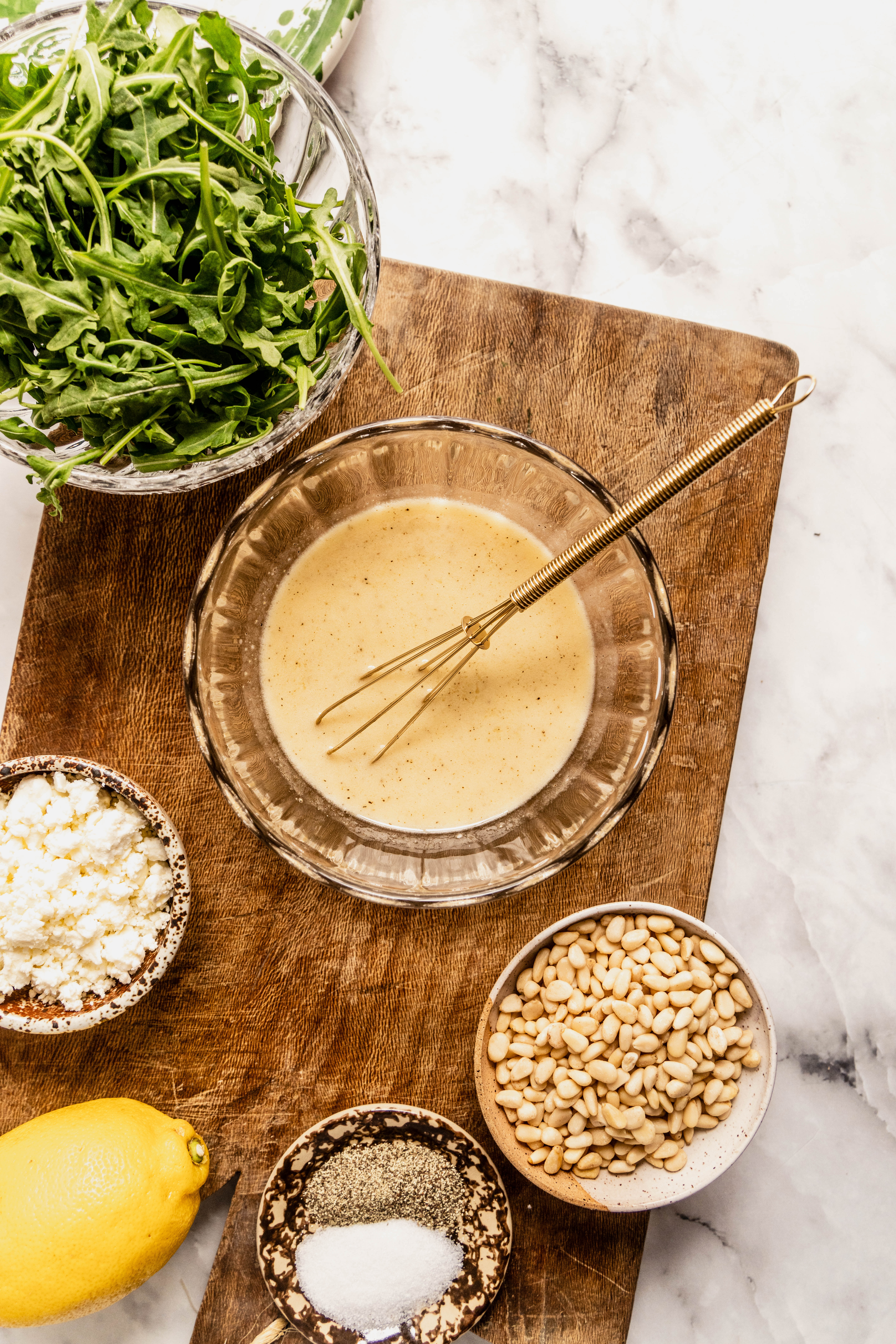 “Overhead view of lemon Dijon vinaigrette being whisked in a glass bowl with arugula, feta cheese, pine nuts, lemon, salt, and pepper on a wooden cutting board, prepared for asparagus orzo salad.”