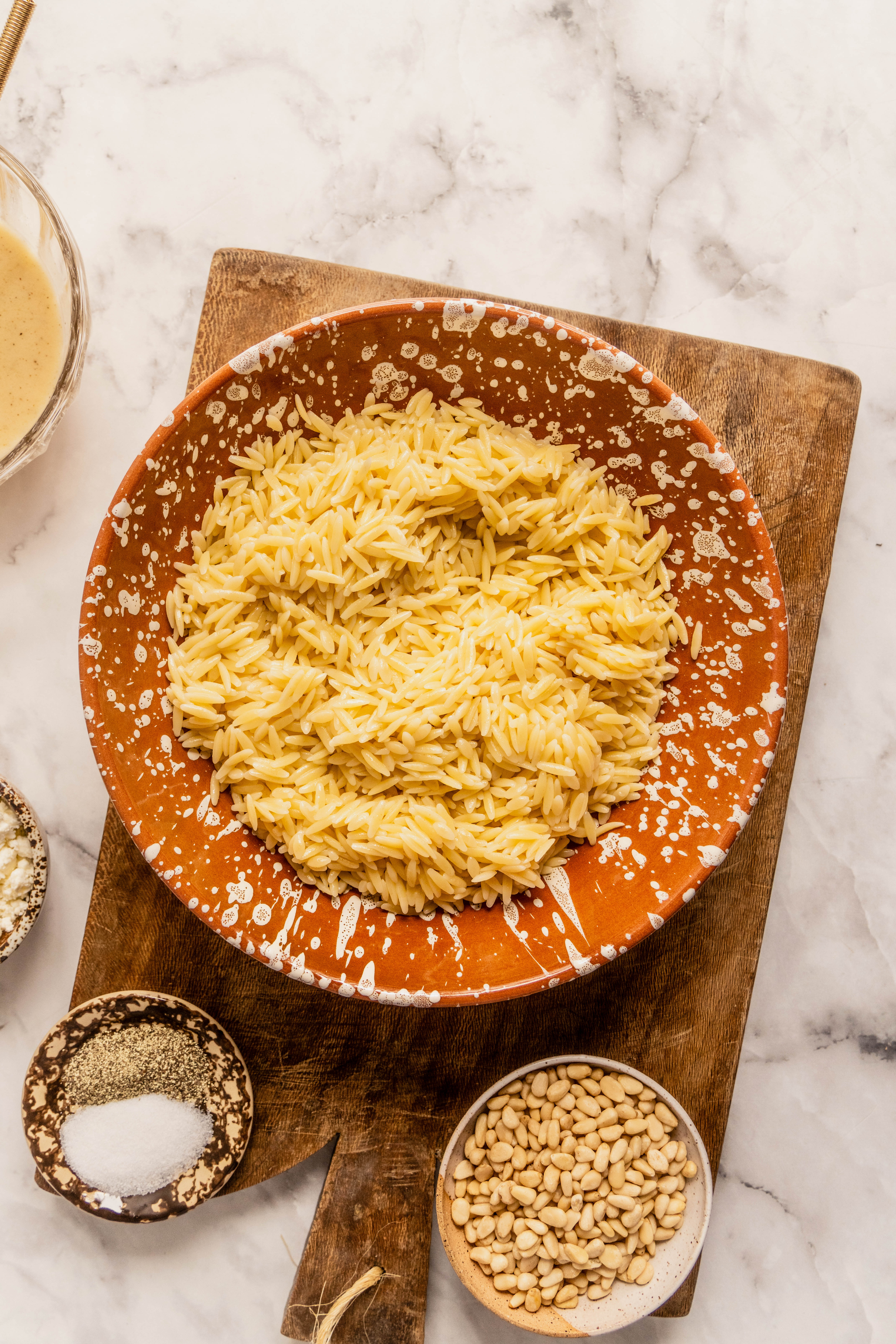 “Overhead view of cooked orzo pasta in a ceramic bowl with pine nuts, salt, and black pepper on a wooden cutting board, prepared for an asparagus orzo salad on a white marble countertop.”