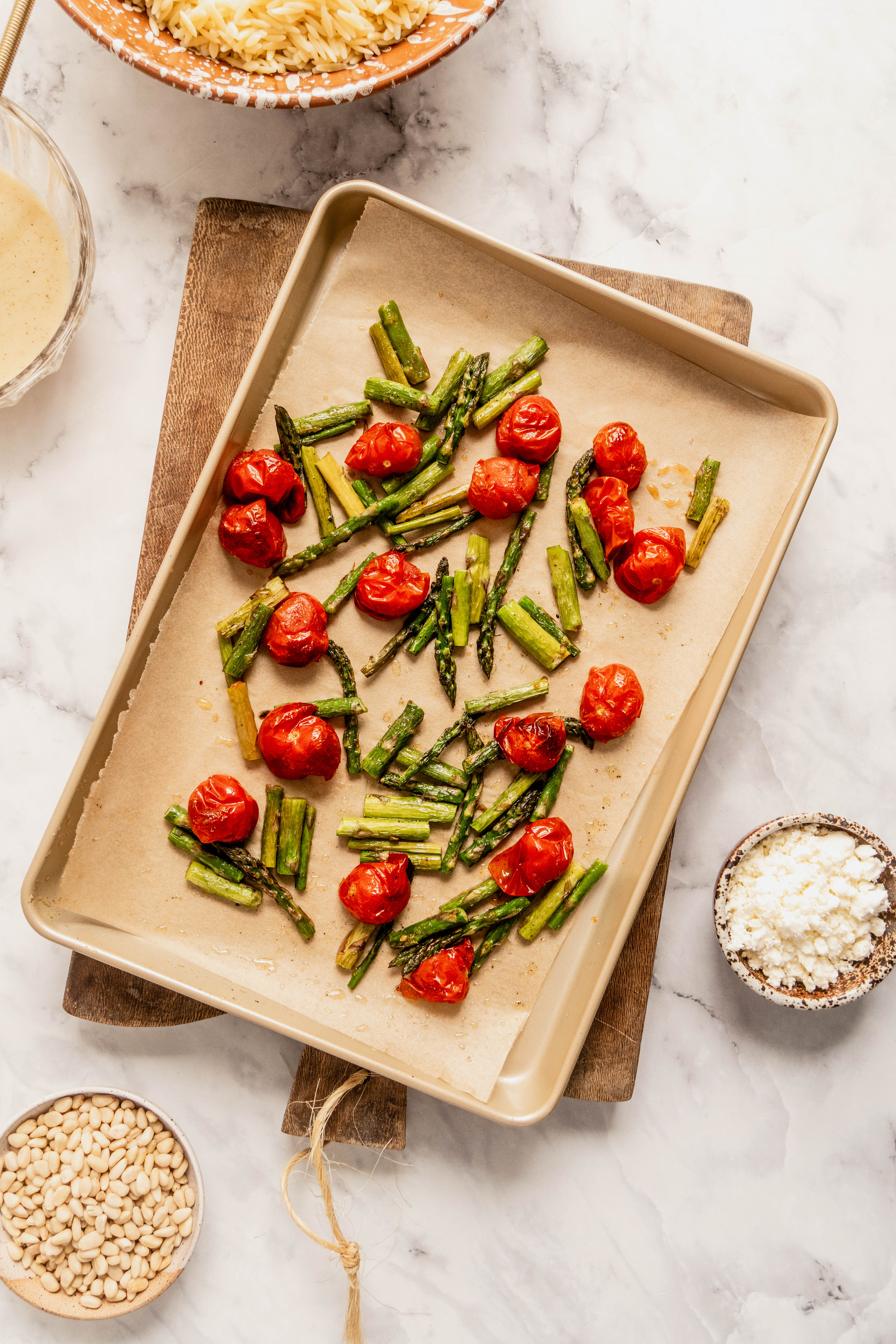“Overhead view of roasted asparagus and blistered cherry tomatoes on a parchment-lined baking sheet, prepared as a component for asparagus orzo salad, with bowls of feta cheese and pine nuts on a white marble surface.”