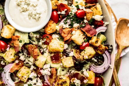 Chopped BLT salad in a large serving bowl topped with crispy bacon pieces, golden croutons, crumbled blue cheese, grape tomatoes, and sliced red onion over chopped lettuce, served with a small bowl of creamy blue cheese dressing on a marble surface with fresh herbs nearby.