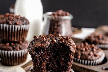 Close-up of a moist Chocolate Zucchini Muffin split open to show a rich, fudgy crumb with melted chocolate chips, styled on vintage book pages with more muffins and a glass bottle of milk softly blurred in the background.