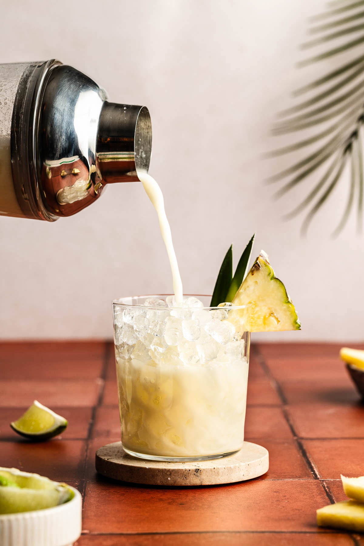 Coconut Water Mocktail being poured from a cocktail shaker into a short glass filled with crushed ice, garnished with a fresh pineapple wedge and pineapple leaves, set on a terracotta tile surface with lime wedges and pineapple slices nearby.