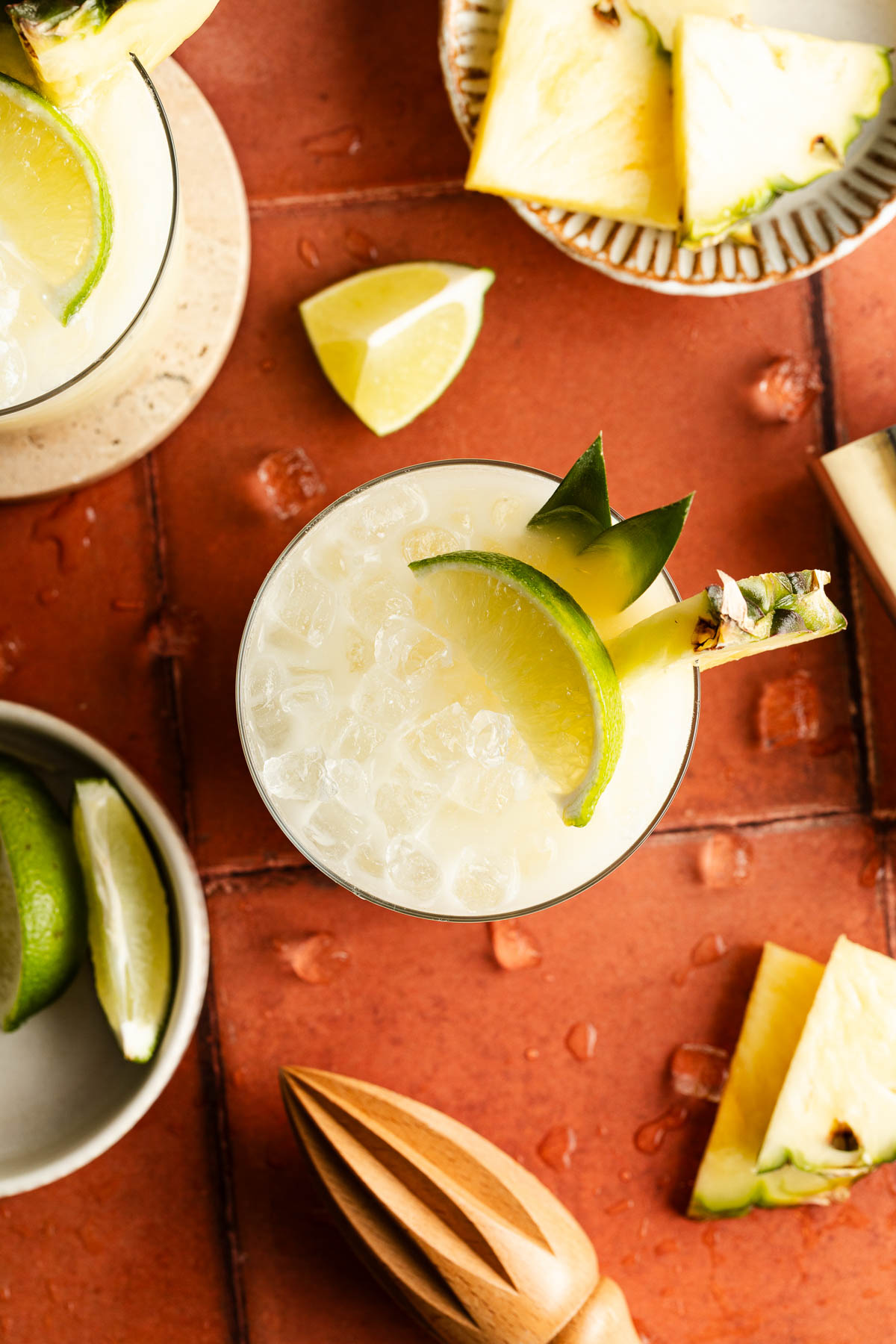Overhead view of a Coconut Water Mocktail in a short glass filled with crushed ice, garnished with a lime wheel and fresh pineapple wedge, sitting on a round stone coaster over warm terracotta tiles with droplets of condensation for a tropical, refreshing feel.