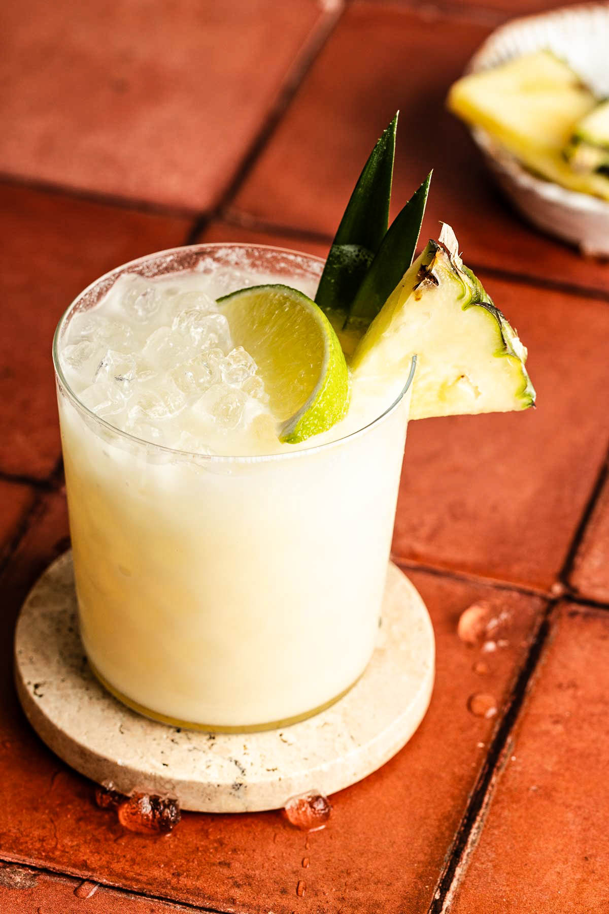 Close-up of a Coconut Water Mocktail in a short glass filled with crushed ice, garnished with a lime wheel and fresh pineapple wedge, sitting on a round stone coaster over warm terracotta tiles with droplets of condensation for a tropical, refreshing feel.