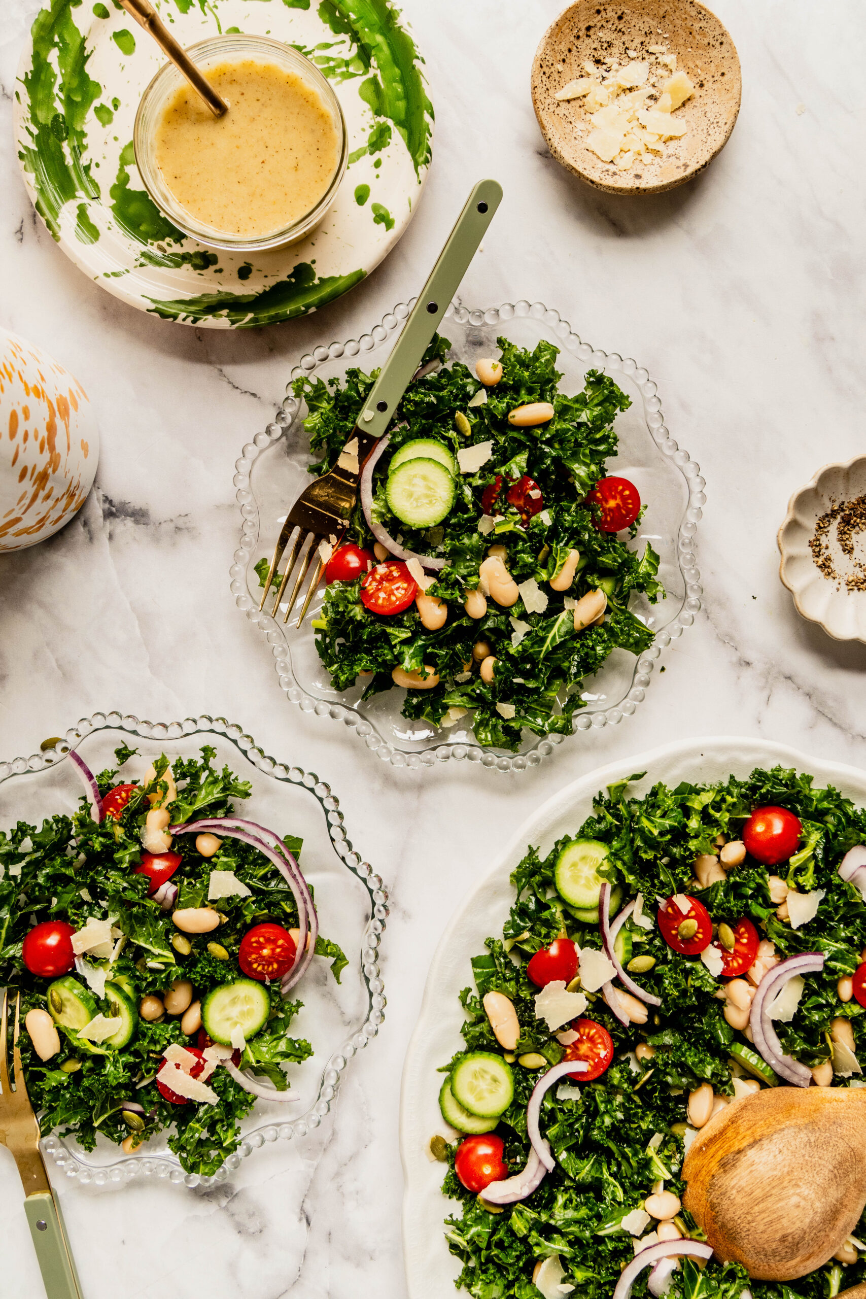 Overhead view of kale and cannellini bean salad served on a large white platter and individual glass plates, topped with cherry tomatoes, sliced cucumbers, red onion, pepitas, and shaved Pecorino, with lemon Dijon dressing nearby on a marble countertop.

