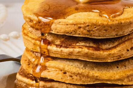 Close-up of gluten free sweet potato pancakes stacked high on a white plate, topped with a pat of melting butter and maple syrup dripping down the fluffy layers in warm natural light.